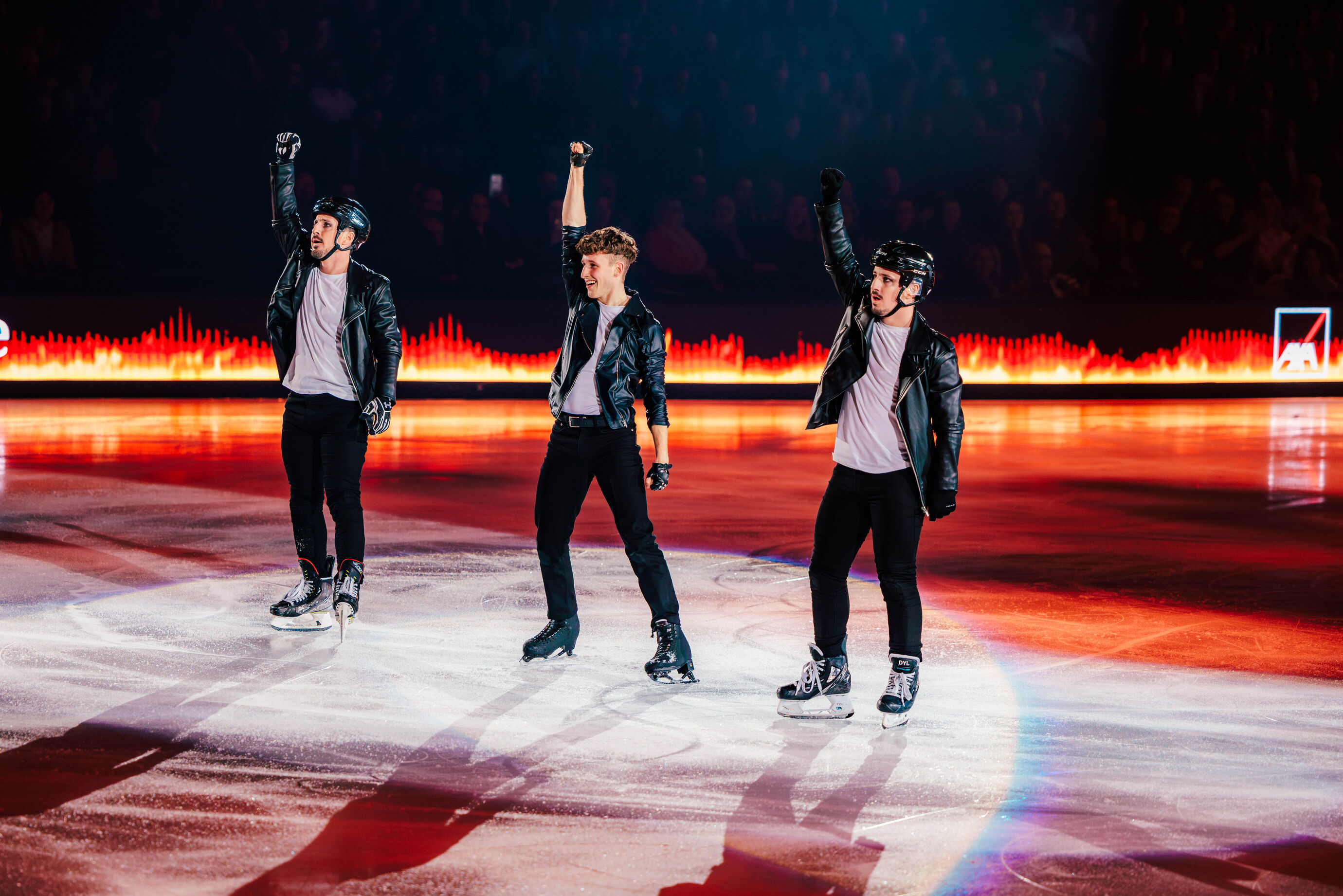 Drei Eiskunstläufer heben synchron die Arme im Scheinwerferlicht.