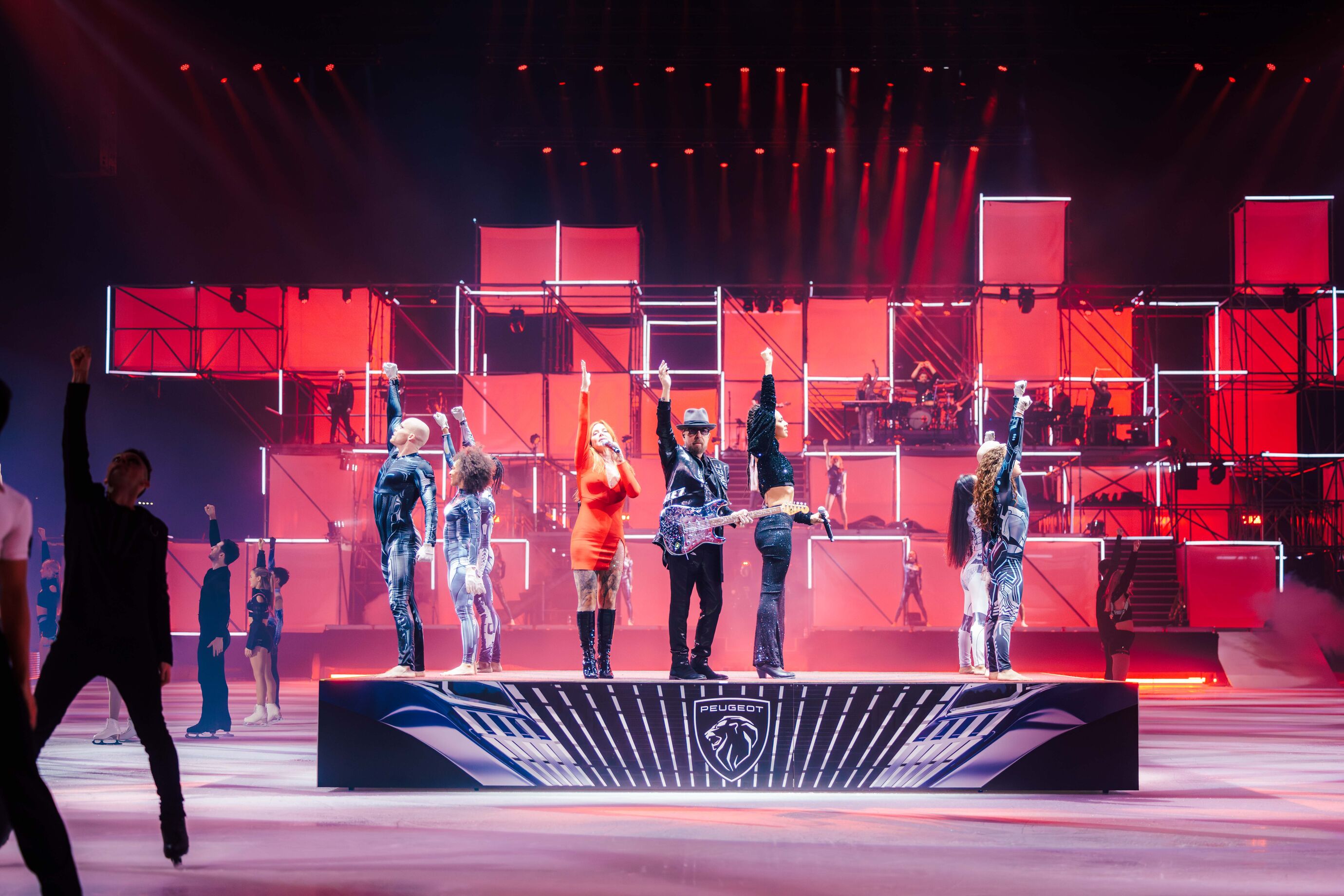 Mehrere Eisläufer und Künstler stehen gemeinsam auf einer Bühne im roten Licht.